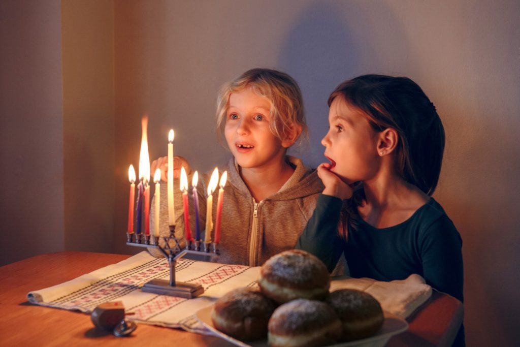 Girls Friends Lighting Candles On Menorah For Traditional Jewish Hanukkah Holiday At Home. Children Celebrating Chanukah Festival Of Lights Together. Dreidel And Sufganiyot Donuts In Plate On Table.